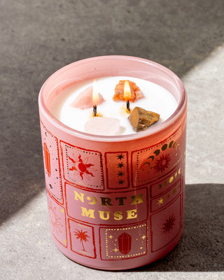 Lit North Muse candle in a pink jar with crystals embedded in the wax and celestial designs on the jar.