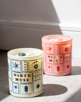 Two crystal-infused scented candles from North Muse placed on a concrete surface. The white jar candle with blue and gold celestial designs is in the foreground, while the pink jar candle with red and gold designs is slightly blurred in the background. Both candles have visible crystals embedded in the wax