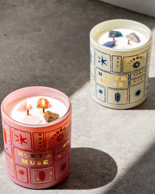 Two crystal-infused scented candles from North Muse, one in a pink jar and the other in a white jar, both with celestial-themed illustrations. The pink candle is lit, showing crystals embedded in the wax, while the white candle in the background is unlit.