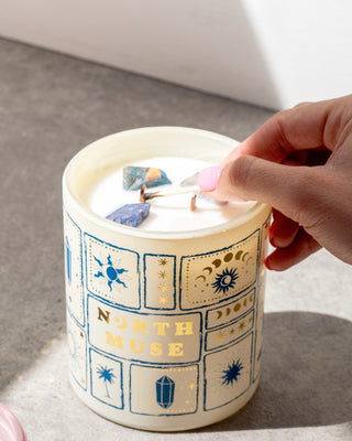 Close-up of a hand with pink-painted nails lighting a candle in a white jar labeled 'North Muse.' The candle has decorative illustrations of celestial symbols and crystals embedded in the wax