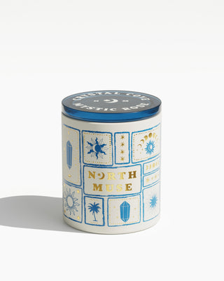 Product image of a North Muse Crystal Cove candle and its packaging. The candle is in a white jar with blue and gold celestial designs and a matching blue lid. The box beside it features the same celestial-themed illustrations and branding, with text indicating it is a 'Crystal & Rock Infused Sea Salt & Sage Scented Soy Candle' weighing 430g (15oz)