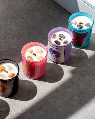 Four North Muse candles in various colors (black, pink, purple, blue) with crystal infusions, arranged on a gray surface with sunlight casting shadows.