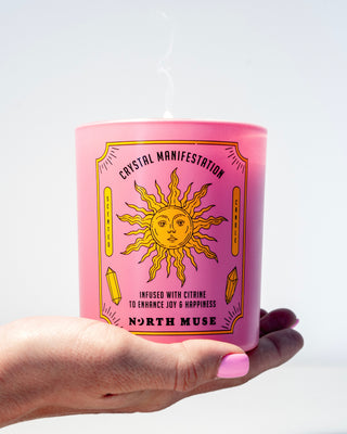 Hand holding a North Muse 'Crystal Manifestation' candle in a pink jar with a sun design, infused with citrine.