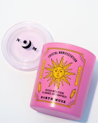 North Muse 'Crystal Manifestation' candle in a pink jar with a sun design, lying on its side next to the lid.