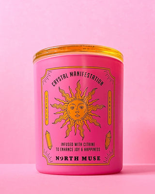 North Muse Crystal Manifestation candle in a pink jar with a sun design, placed against a matching pink background.