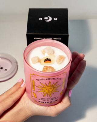 Hands holding a lit North Muse Crystal Manifestation candle in a pink jar with a sun design, with a black box in the background.