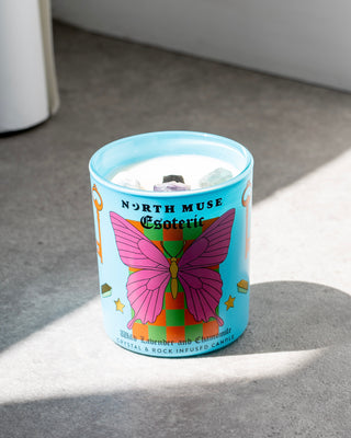 North Muse Esoteric candle in a blue jar with a pink butterfly design, infused with crystals, placed on a gray surface.