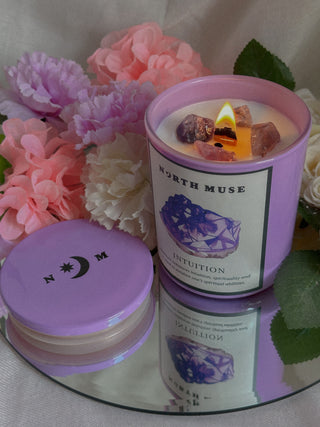 A lit North Muse "Intuition" candle in a light purple jar, surrounded by soft pink and white flowers. The candle sits on a reflective surface with its lid beside it, creating a serene and mystical ambiance.