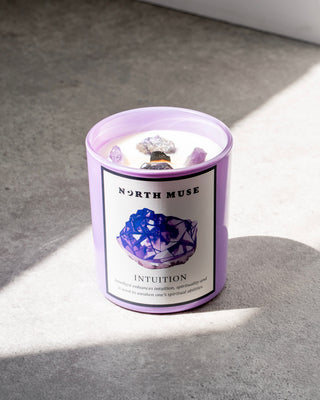 A North Muse candle in a light purple jar labeled "Intuition," featuring a detailed illustration of an amethyst crystal cluster. The candle is placed on a gray surface with sunlight illuminating part of the scene.