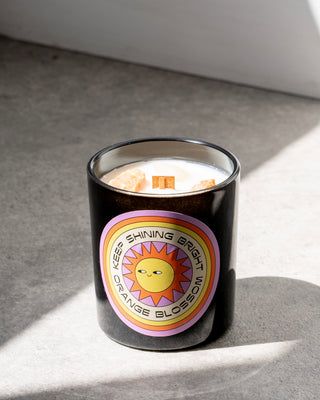 North Muse candle in a black jar with a "Keep Shining Bright" label featuring a sun and "Orange Blossom" text, placed on a concrete surface.