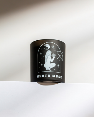 North Muse black jar candle with white celestial illustrations, placed on a white surface. Brand name displayed at the bottom.