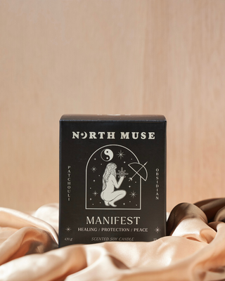 Black North Muse candle box labeled 'Manifest' with the words 'Healing / Protection / Peace' and 'Patchouli' and 'Obsidian' written on the sides. The box features a white illustration of a kneeling figure holding a star, surrounded by celestial symbols, including a yin-yang. The box is placed on soft, light-colored fabric against a neutral background.