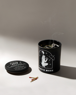 North Muse 'Manifest' candle in a black jar with a celestial design, featuring a kneeling figure holding a star. The candle has just been extinguished, with wisps of smoke rising from the wick. The candle lid, which is black with white text, is placed beside the jar, along with a few burnt matches.