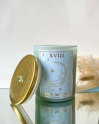 North Muse 'The Moon' candle with gold lid placed on a reflective surface, featuring a celestial crescent moon design in gold, set against a neutral background.