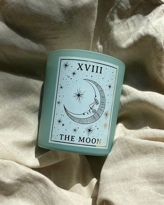 North Muse 'The Moon' candle with celestial crescent moon design, lying on soft beige fabric, bathed in natural light.