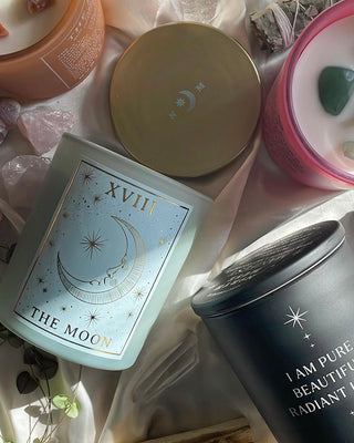 Collection of North Muse candles including 'The Moon' with celestial crescent moon design, displayed with a gold lid and surrounding crystals on soft satin fabric.