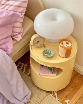 Cozy bedroom nightstand setup featuring a white mushroom lamp, beige retro clock, ceramic jewelry dish, and North Muse 'The Sun' candle in an orange jar. The nightstand holds a pastel pink book, placed beside a bed with lilac bedding.