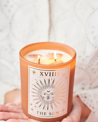 Person holding a burning North Muse 'The Sun' candle with a celestial sunburst design, set against a soft, white fabric background.