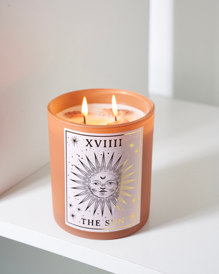 North Muse 'The Sun' candle with celestial sunburst design, burning with two wicks, placed on a white shelf against a light background.