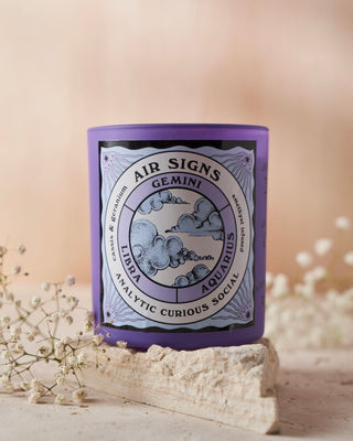 North Muse 'Air Signs' candle in a purple jar featuring Gemini, Libra, Aquarius zodiac signs, placed on a stone with floral accents.