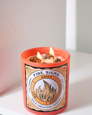 Lit North Muse Fire Signs candle in an orange jar featuring zodiac signs Aries, Leo, and Sagittarius, with crystal infusions.