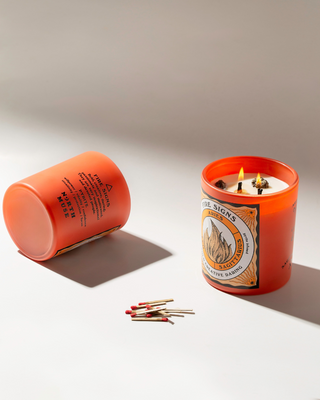 Lit North Muse Fire Signs candle in an orange jar with a second candle lying on its side, next to a few matches on a white surface.