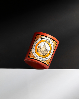 North Muse Fire Signs candle in an orange jar featuring zodiac signs Aries, Leo, and Sagittarius, photographed against a black background.