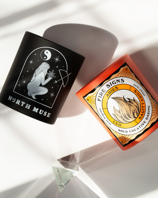 North Muse black candle with celestial design and Fire Signs orange candle, both lying on their sides with light reflections on a white surface.