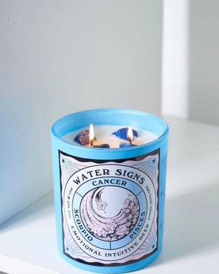 North Muse 'Water Signs' candle in a light blue jar with a wave illustration and zodiac signs Cancer, Scorpio, and Pisces, labeled 'Emotional, Intuitive, Deep.' The candle is lit, with two flames and visible blue crystals embedded in the wax. The candle is placed on a white surface against a neutral background.