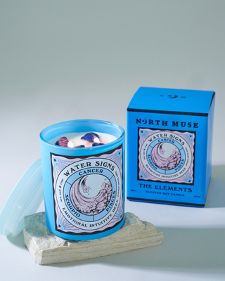 North Muse 'Water Signs' candle in a light blue jar with a wave illustration and zodiac signs Cancer, Scorpio, and Pisces labeled 'Emotional, Intuitive, Deep.' The candle is lit, with visible crystals embedded in the wax. The jar is placed on a stone slab, with the matching blue box in the background, labeled 'The Elements' and 'Scented Soy Candle.