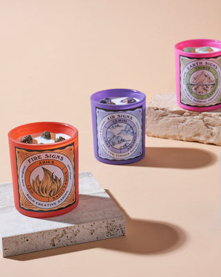 Three North Muse candles representing Fire Signs, Air Signs, and Earth Signs, in orange, purple, and pink jars, arranged on different surfaces.