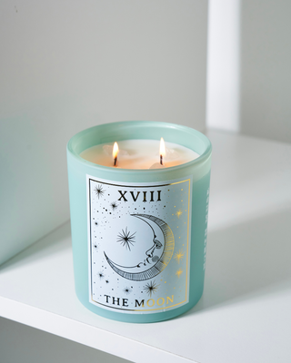 North Muse 'The Moon' candle with a celestial design, burning with two wicks, placed on a white shelf.