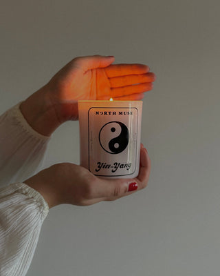 Hands holding a lit North Muse Yin-Yang candle with a glowing flame. The candle features a Yin-Yang symbol and text 'North Muse' on the label, creating a serene and calming ambiance.