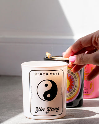 Hand lighting a North Muse Yin-Yang candle with a match, with the candle featuring a black Yin-Yang symbol and text on the label. Other colorful candles are visible in the background.