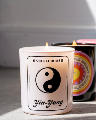 North Muse Yin-Yang candle in a white jar with black yin-yang symbol, featuring a burning wick. The candle is labeled 'Cherry Blossom' and 'Rose Quartz,' with another lit candle visible in the background.