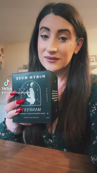 Woman with long dark hair holding a North Muse 'Manifest' candle box, featuring celestial designs. TikTok username '@curiousmagpiereadings' visible.